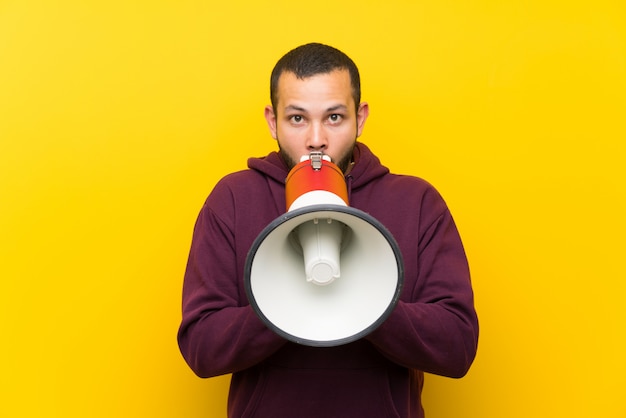 Kolumbianischer Mann mit Sweatshirt über gelber Wand schreiend durch ein Megaphon