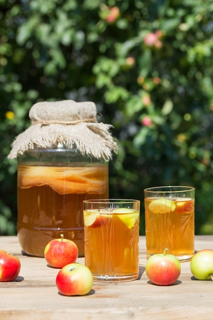 Kombucha fermentada bebe em uma jarra de vidro e dois copos com uma bebida com maçãs em primeiro plano, em um dia ensolarado de verão no jardim em uma mesa de madeira. Estilo rústico.