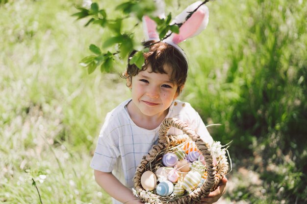 Foto komischer junge mit eierkorb und kaninchenohren auf der ostereierjagd im sonnigen frühlingsgarten