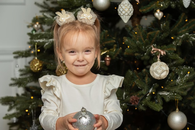 Komisches kleines Mädchen mit einem Weihnachtsball auf dem Hintergrund eines Weihnachtenbaums