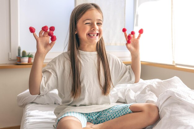 Komisches kleines Mädchen mit Himbeeren an den Fingern im Bett am Morgen