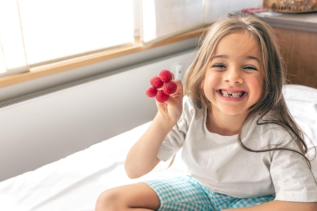 Komisches kleines Mädchen mit Himbeeren an den Fingern im Bett am Morgen