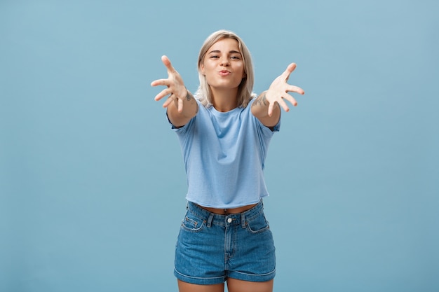 Komm zu mama. porträt der freundlichen freudigen und enthusiastischen kaukasischen studentin im trendigen outfit, das hände zu den lippen zieht und faltet, um zu umarmen und leidenschaftlichen kuss zu geben, der über blaue wand lächelt