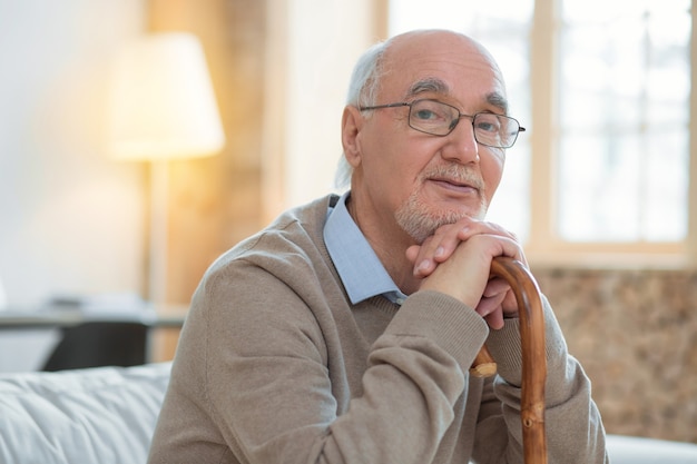 Komm zu mir. Meditativer attraktiver älterer Mann, der sich auf Zuckerrohr stützt, während er Brille trägt und Kamera betrachtet