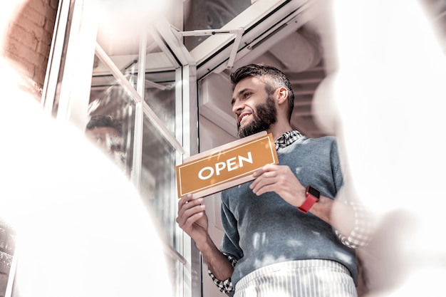 Komm zu uns. niedriger winkel eines netten angenehmen mannes, der nahe der kaffeetür steht, während er ein türschild hält