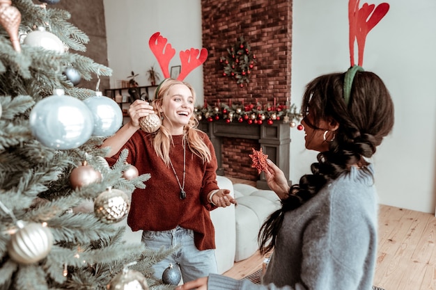 Kommende Ferien. Zufriedenes blondes Mädchen, das ihrem Freund abgerundetes Spielzeug zeigt, während es im dekorierten stilvollen Wohnzimmer-Weihnachtskonzept bleibt