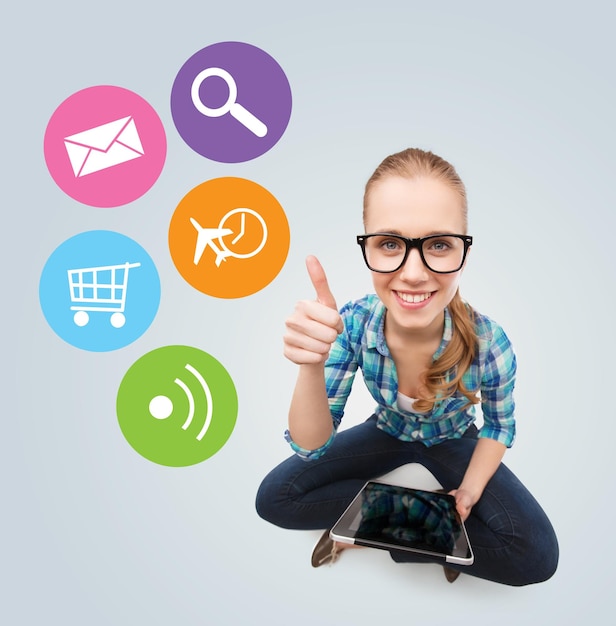Kommunikations-, Technologie-, Internet- und Personenkonzept - lächelndes Teenager-Mädchen mit Brille, das auf dem Boden sitzt und Tablet-PC-Computer über grauem Hintergrund mit bunten Symbolen hält