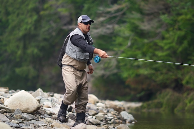 Kompetente Fischer in Mütze, Brille und wasserdichtem Outfit, die mit Rute zwischen Bergen angeln. Mann, der im Fluss steht und Fisch am Haken fängt.