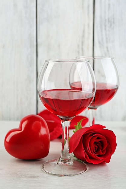 Komposition mit Rotwein im Glas, roten Rosen und dekorativem Herzen auf farbigem Holzhintergrund