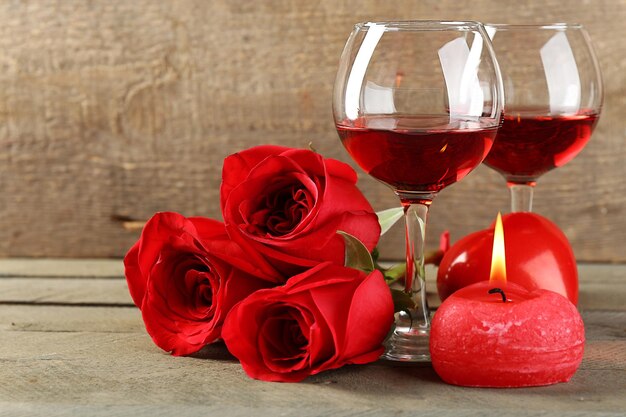Komposition mit Rotwein in Gläsern, roten Rosen und dekorativem Herzen auf Holzuntergrund