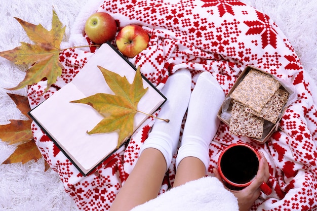 Komposition mit warmem Plaid, Buch, Tasse heißem Getränk und weiblichen Beinen, auf farbigem Teppichhintergrund