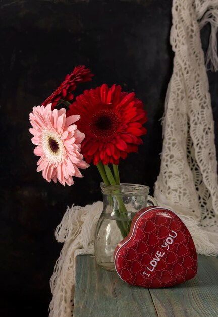 Komposition zum Valentinstag, Frauentag oder Geburtstag. Romantischer Blumenstrauß aus Gerbera und eine Schachtel Süßigkeiten in Form eines Herzens auf einem Holztisch in der Nähe.