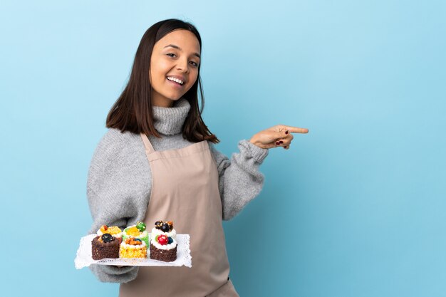 Konditor hält einen großen Kuchen über der blauen Wand und zeigt mit dem Finger zur Seite