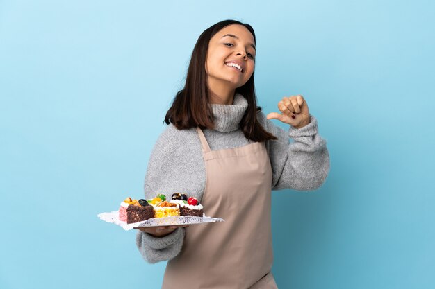 Konditor hält einen großen Kuchen über isolierten blauen Raum stolz und selbstzufrieden