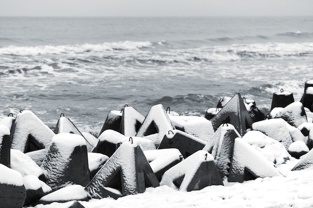 Konkrete Wellenbrecher vor dem Hintergrund des Wintermeeres mit Schnee bedeckt. Küstenschutz im Schnee. Verschneite bewölkte Meereslandschaft.
