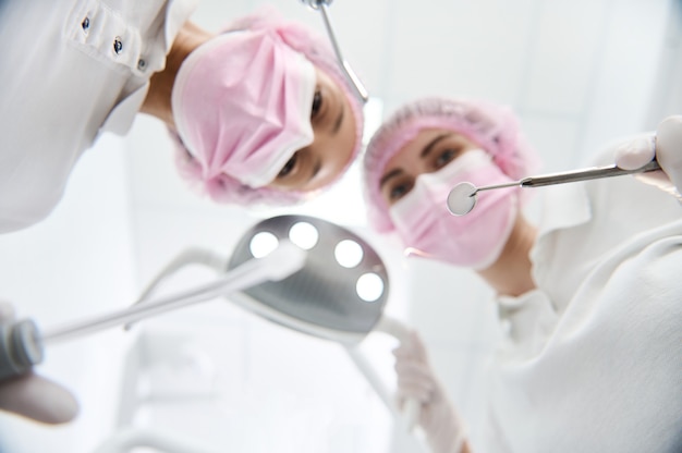 Konzentrieren Sie sich auf einen Zahnspiegel aus Edelstahl in der Hand eines verschwommenen weiblichen Chirurgen, der ein Licht neben dem Assistenten mit Speichelsauger und zahnärztlicher Ausrüstung zur Behandlung menschlicher Zähne einstellt
