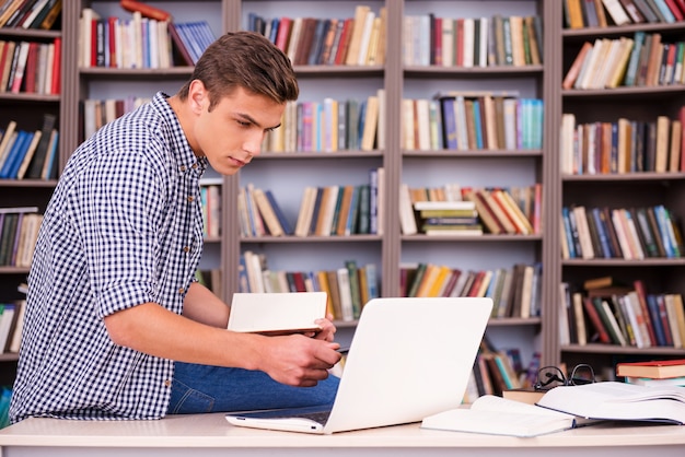 Konzentriert auf das Studium. Selbstbewusster junger Mann, der Notizblock hält und Laptop beim Sitzen am Schreibtisch und in der Schrift des Bücherregals betrachtet