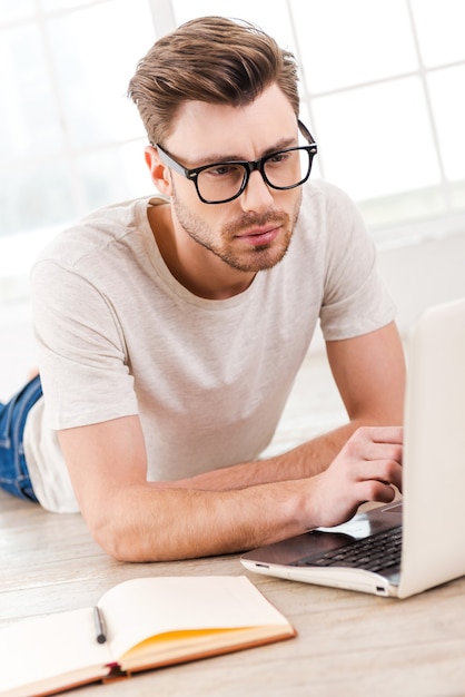 Konzentriert auf die Arbeit. Nachdenklicher junger Mann, der am Laptop arbeitet, während er zu Hause auf dem Boden sitzt