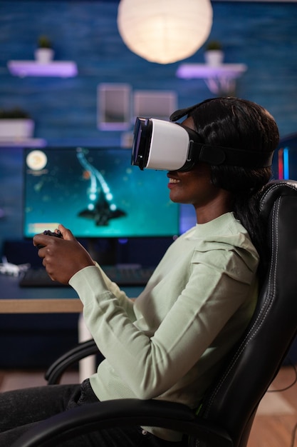 Konzentrierte afroamerikanische Streamerin mit Virtual-Reality-Headset, die während des Online-Turniers Weltraum-Shooter-Videospiele mit Gaming-Controller spielt. RGB-Computerausrüstung. Cyberspace