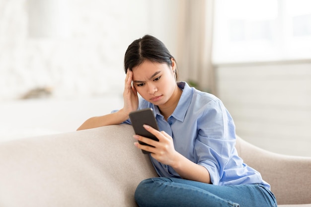 Konzentrierte asiatische Frau, die mit dem Handy auf der Couch sitzt