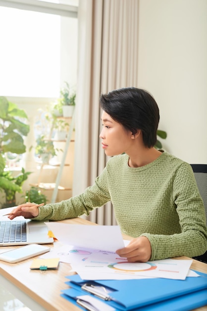 Konzentrierte asiatische Frau sitzt am Tisch und arbeitet am Laptop und macht sich Notizen im Notizbuch im Heimbüro