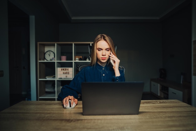 Konzentrierte Dame in einem blauen Hemd sitzt zu Hause an einem Tisch mit einem Laptop und arbeitet aus der Ferne