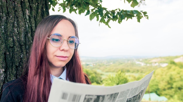 Konzentrierte elegante Dame, die Zeitung im Park liest Fokussierte junge Unternehmerin mit langem Haar in formeller Kleidung und Brille, die in der Nähe von Bäumen steht und interessiert Zeitung liest