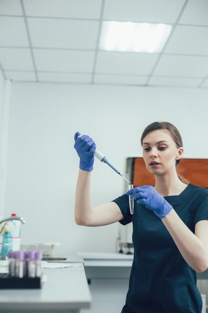 Konzentrierte Frau in Gummihandschuhen, die im Labor steht und ein Reagenzglas mit einer Pipette verwendet