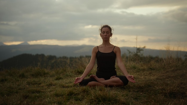 Konzentrierte Frau meditiert in den Bergen bei Sonnenuntergang. Entspanntes Mädchen sitzt im Lotussitz auf dem Boden im Freien. Athletische Dame praktiziert Yoga-Pose in der Sommerlandschaft