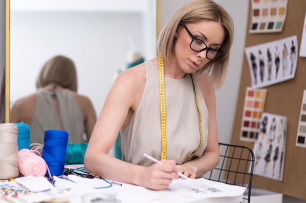 Foto konzentrierte frau mit maßband arbeitet mit kleidungskizze gegen spiegel im modestudio