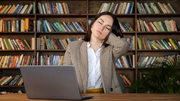 Konzentrierte Freiberuflerin tippt auf grauem Laptop und streckt den Nacken sitzend auf einem Stuhl gegen große Holzregale mit farbigen Büchern