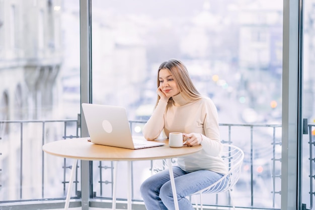 Konzentrierte junge Geschäftsfrau oder Studentin, die auf einen Laptop schaut, der eine Kaffeetasse hält und eine ernsthafte Frau studiert, die mit einem Computer arbeitet oder studiert, der online recherchiert oder sich auf die Prüfung vorbereitet