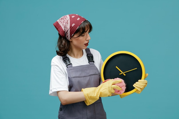 Konzentrierte junge Putzfrau mit einheitlichem Kopftuch und Gummihandschuhen, die die Uhr hält und mit Staubtuch isoliert auf blauem Hintergrund abwischt