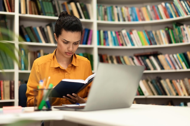 Konzentrierte Mischlingsfrau, die ein Buch liest, das in der Bibliothek mit Laptop-PC und Studienmaterialien sitzt, kostenloser Kopienraum