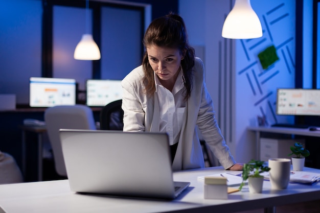 Konzentrierte müde Geschäftsfrau, die spät in der Nacht E-Mails auf einem Laptop liest, der in der Nähe des Schreibtisches in einem Start-up-Unternehmen steht
