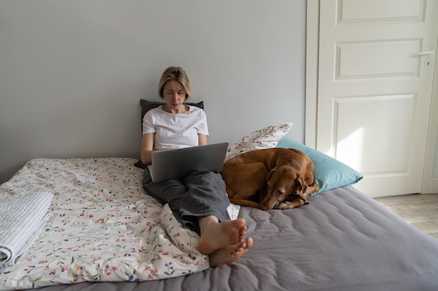 Foto konzentrierte, nachdenkliche frau mit hund sitzt auf dem bett und streckt die beine mit einem laptop für die online-bildung