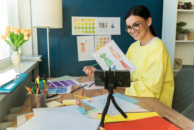 Konzentrierte professionelle Designerin, die Skizzen von neuem Design für stilvolle Kleidung zeigt, während sie im eigenen Atelier sitzt und per Videoanruf am Smartphone chattet