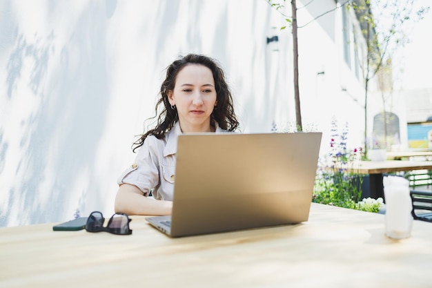 Konzentrierte Studentin schreibt auf dem Laptop, während sie mit einer Tasse im Café studiert. Nachdenkliche Freiberuflerin notiert Informationen für die Projektplanung und erledigt Remote-Arbeit per Laptop