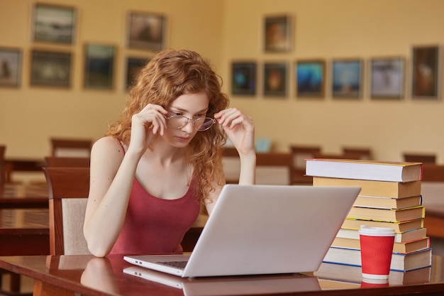 Konzentrierte Studentin studiert in der Schulbibliothek, benutzt einen Laptop und sucht nach Informationen, nutzt das drahtlose Internet, hält die Hände auf dem Brillengestell und schaut auf den Computer. Bildungskonzept.