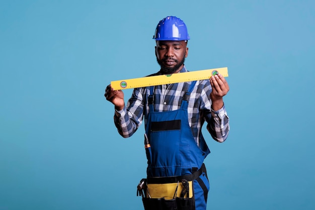 Konzentrierter afrikanisch-amerikanischer Bauarbeiter in Uniform mit Richtmaschine auf blauem Hintergrund. Erfahrener Baumeister mit Arbeitswerkzeugen in Gürtel und Schutzhelm, Studioaufnahme.