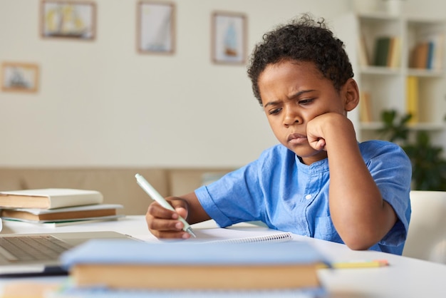 Konzentrierter afroamerikanischer Schuljunge im blauen Hemd, der am Schreibtisch sitzt und zu Hause Unterricht lernt