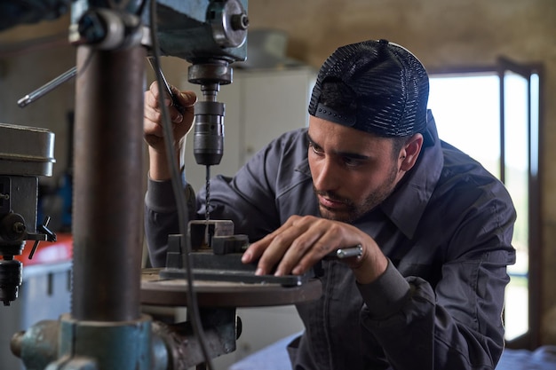 Konzentrierter bärtiger männlicher Techniker in grauer Uniform und Mütze, der während des Arbeitstages in der Reparaturwerkstatt mit einem Vertikalbohrer Metalldetails präzise bohrt