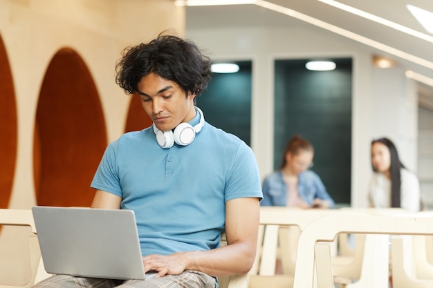 Konzentrierter dunkelhäutiger Student, der auf Stuhl in Bibliothek sitzt und Laptop benutzt, während er an Universitätsprojekt arbeitet