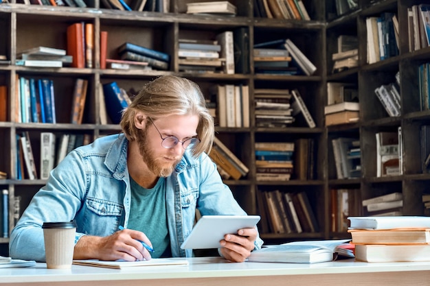 Foto konzentrierter, ernsthafter mann, schüler, lehrer, der einen digitalen tablet-computer benutzt, auf den bildschirm schaut, videos ansieht, notizen macht, online-app-kurs lernt, im internet lernt, am bibliotheksschalter sitzt, selbstbildungskonzept