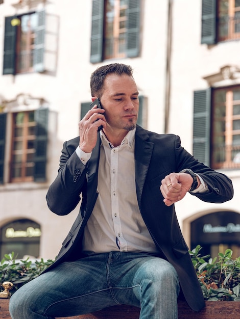 Konzentrierter erwachsener bärtiger männlicher Unternehmer mit dunklen Haaren in schlauer Freizeitkleidung, der auf einer Bank sitzt und auf ein Meeting wartet, telefoniert mit seinem Smartphone und überprüft die Zeit auf seiner Armbanduhr