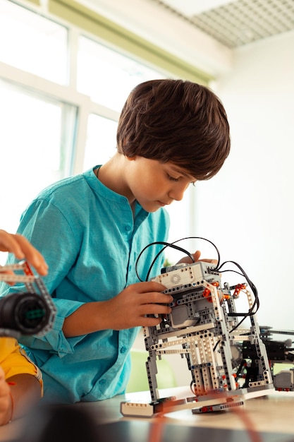 Konzentrierter Grundschüler, der im Klassenzimmer neben seinem Schreibtisch steht und sein kompliziertes Wissenschaftsprojekt aus Bausatz überprüft