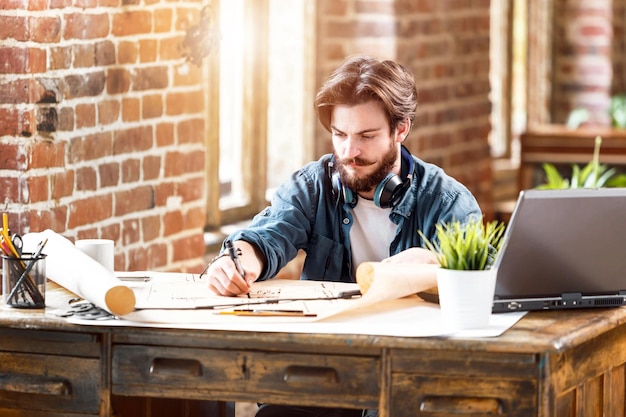 Konzentrierter junger Architekt, der mit Blaupause arbeitet, während er mit Laptop in einem sonnigen Büro im Dachgeschoss sitzt