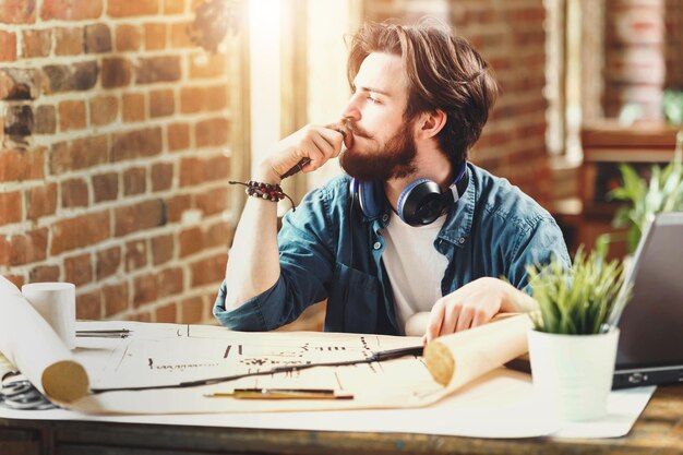 Konzentrierter junger Architekt, der zum Fenster blickt und über einen neuen Bauplan nachdenkt, während er mit einem Laptop in einem sonnigen Büro im Loft sitzt