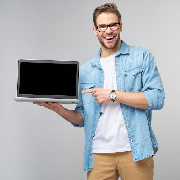 Konzentrierter junger bärtiger Mann, der eine Brille trägt, gekleidet in das Jeanshemd, das Laptop hält