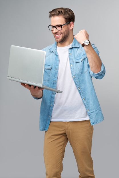 Konzentrierter junger bärtiger Mann, der eine Brille trägt, gekleidet in das Jeanshemd, das Laptop hält